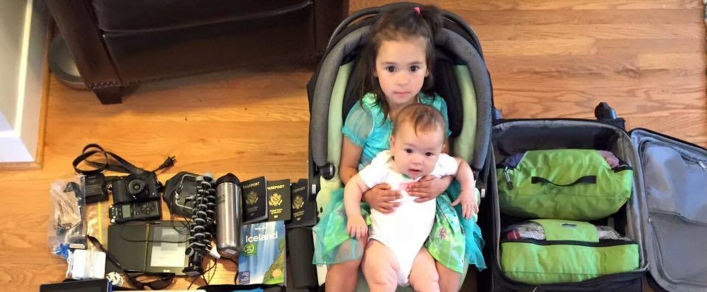 Chloe holding baby Jasmine in a car seat, surrounded by three months of travel gear laid out on the floor: packing cubes, boots, an Iceland guidebook, passports
