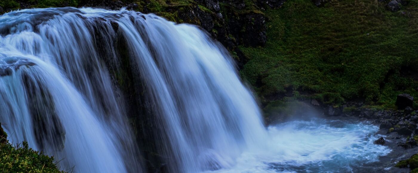 A wide waterfall dropping over a dark ledge into a green gorge in Iceland